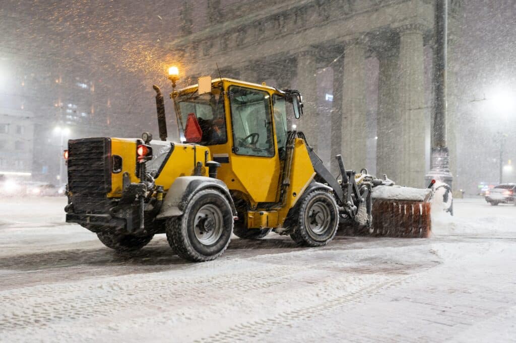 Winter service vehicles. Big yellow snow plow tractor working in city at night in heavy snowfall